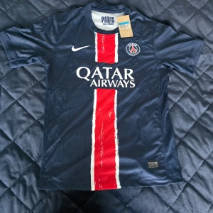 PSG matchtröja Nike marinblå röd - Paris Saint-Germain fotbollströja från Nike i marinblått med en bred röd rand och vita detaljer. PSG-logga på bröstet, Qatar Airways-tryck och klassisk rund hals. Perfekt för dig som vill visa ditt stöd för PSG på match eller träning.