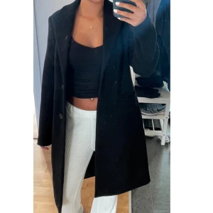 Svart lång kappa med knappar - Säljer en svart, stilren kappa med klassisk krage och två stora knappar framtill. Jackan har en rak och längre passform som ger en snygg siluett. Perfekt för dig som gillar enkel och clean stil. Materialet känns mjukt och har en lite tjockare känsla.