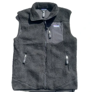 Svart fleeceväst från Patagonia - Mysig svart fleeceväst från Patagonia med hög krage och dragkedja framtill. Västen har tre fickor med dragkedja, varav en på bröstet med logotyp. Perfekt för lager-på-lager och outdoor vibes.