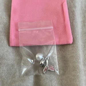 Pandora berlock med pärla, rosa stenar och en fena - Säljer en söt Pandora berlock i silverfärg med en glittrig valfena dekorerad med rosa stenar och en vit pärla. Perfekt att hänga på ditt armband för en personlig touch. Kommer i en liten plastpåse med rosa tygpåse.