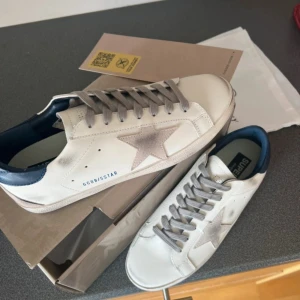 Golden Goose Superstar sneakers vit/grå - Golden Goose Superstar sneakers i vitt skinn med grå stjärndetalj på sidan, grå snörning och mörkblå häl. Klassisk låg modell med rund tå och platt sula. Snyggt slitna detaljer för en cool, avslappnad look. Kommer med originalkartong och dustbag.