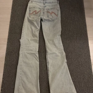 Ljusblå bootcut jeans från Nelly - Snygga ljusblå bootcut jeans från Nelly med slitna hål på knäna och coola broderade detaljer i rosa på bakfickorna. Jeansen har klassisk femficksdesign och är tillverkade i mjukt denimtyg. Perfekta för dig som gillar en avslappnad och trendig look.