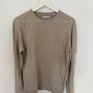 Beige stickad tröja från Jack & Jones - En stilren beige stickad tröja från Jack & Jones i storlek XS. Tröjan har rund halsringning, långa ärmar och ribbade muddar vid ärmslut och nederkant. Perfekt för lager-på-lager och enkel att matcha med jeans eller chinos.
