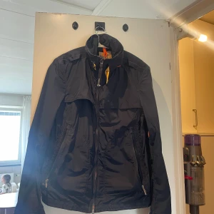 Svart vindjacka från Parajumpers - Svart vindjacka från Parajumpers med hög krage, orange detaljer och dragkedjor. Jackan har flera fickor med dragkedja och en cool gul rem vid halsen. Perfekt för dig som vill ha en snygg och funktionell jacka till vardags. Har någon liten skråma baktill men inget som förstör funktionen 