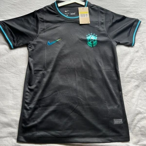 Svart Brasilien fotbollströja Nike - Snygg svart Brasilien fotbollströja från Nike med korta ärmar och turkosa detaljer vid krage och ärmslut. Tröjan har Brasilien-emblem på bröstet och Nike-logga i turkos. Tillverkad i lätt och funktionellt material som andas.