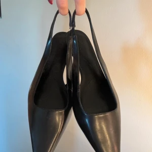 Svarta pumps med låg klack - Stilrena svarta pumps i skinn med spetsig tå och låg klack. Skorna har öppen häl med smal rem som ger en elegant och modern look. Perfekta för dig som gillar klassiska och minimalistiska skor med en twist.