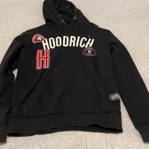 Svart hoodie från Hoodrich med patchar - Svart hoodie från Hoodrich i storlek M med coola broderade patchar och stor vit logga framtill. Tröjan har huva med snörning och långa ärmar. Materialet är mjukt och känns som polyester, perfekt för chill dagar. Snygga detaljer med röd och vit text samt Hoodrich-märke på sidan.