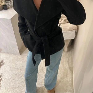 Svart kappa med bälte från vero moda  - Snygg svart kappa med bred krage och knytbälte i midjan. Jackan har två stora fickor framtill och stängs med knappar. Den har en klassisk, stilren look och är långärmad med en normal passform. Perfekt för dig som gillar enkel och clean stil.