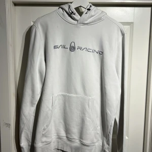 Vit hoodie från Sail Racing, storlek L - Vit hoodie från Sail Racing med broderad logga på bröstet. Klassisk känguruficka framtill, SPARSAMNR ANVÄND.snörning i huvan och mjukt material på insidan. Perfekt för dig som gillar stilren och sportig look.PASAR ÄVEN M