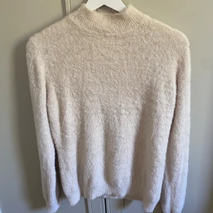Ljusbeige fluffig tröja  - Supermjuk och fluffig ljusbeige tröja från Phi Phi Star med hög hals och långärmad passform. Tröjan är stickad och har en clean look utan mönster eller detaljer, perfekt för dig som gillar mysiga vibes och enkel stil.