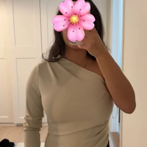 Beige oneshoulder topp Gina Tricot - Säljer en stilren beige oneshoulder topp från Gina Tricot i storlek S. Toppen har lång ärm på ena sidan och ärmlös på andra, med en tight passform och mjukt stretchigt material. Perfekt för dig som gillar minimalistisk och trendig stil.