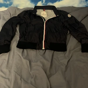 Svart vindjacka från Moncler - Svart vindjacka från Moncler med klassisk krage och ribbade muddar. Jackan har en snygg dragkedja med röd, vit och blå detalj samt Moncler-logga på ärmen. Tillverkad i polyester och har en normal passform. Perfekt för dig som vill ha en stilren och sportig look.