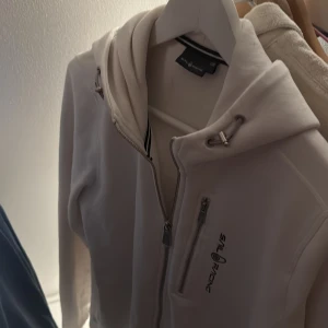 Vit hoodie från Sail Racing med dragkedja - Vit hoodie från Sail Racing med hel dragkedja framtill, snörning i huvan och en bröstficka med dragkedja. Stort Sail Racing-tryck på bröstet. Skön och sportig modell i mjukt material, perfekt för chill eller aktivitet.