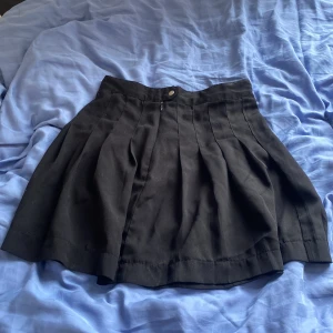 Svart plisserad kjol från Lindex - Svart kortkjol från Lindex med snygga plisserade veck och hög midja. Kjolen har dragkedja och knapp framtill för enkel stängning. Perfekt för dig som gillar en klassisk och stilren look.