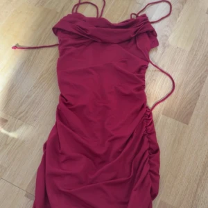Röd kort klänning med smala axelband - Snygg röd kort klänning med smala axelband och rynkade sidor. Klänningen har en draperad urringning och figurnära passform, perfekt för dig som vill sticka ut. Materialet känns mjukt och följsamt mot kroppen.