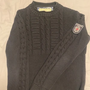 Svart kabelstickad tröja från Morris - Kabelstickad svart tröja från Morris med rund hals och snygga detaljer på axlar och ärmar. Tröjan har ett Morris-märke på ena ärmen och ribbade muddar. 