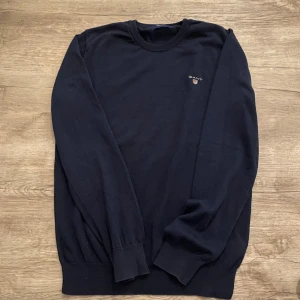 Mörkblå stickad tröja från GANT - Klassisk mörkblå stickad tröja från GANT med rund halsringning och broderad logga på bröstet. Tröjan har ribbade muddar vid ärmslut och nederkant, perfekt för en stilren och enkel look. Kan sänka priset om ni köper mer men kan även sänka annars om ni köper den här!!?