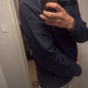 Marinblå vindjacka från J.Lindeberg - Snygg marinblå vindjacka från J.Lindeberg med raka linjer, ståkrage och tryckknappar. Jackan har två bröstfickor med lock och två sidofickor. Materialet är slätt och lätt, perfekt för vår och höst. Diskret logga på ärmen ger en clean look.