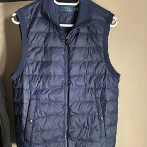 Mörkblå väst från Polo Ralph Lauren - Snygg mörkblå dunväst från Polo Ralph Lauren med quiltad framsida, broderad logga på bröstet och två fickor med knappstängning. Västen har dragkedja framtill och en mjuk, enfärgad baksida. Perfekt för lager-på-lager under höst och vinter. Storlek XS men kan även passa S. Helt ny, aldrig använd. 