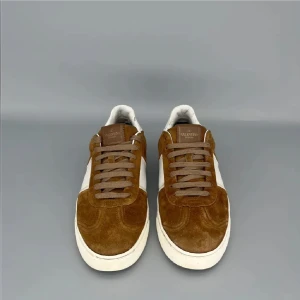 Valentino Garavani bruna sneakers mocka - Snygga sneakers från Valentino Garavani i brun mocka med vita skinnpaneler och coola nitar på hälen. Skorna har rund tå, platt sula och klassisk snörning. Perfekta för dig som vill ha en stilren men ändå unik look.