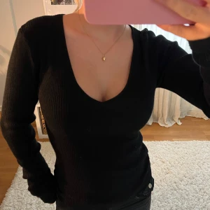 Svart ribbad långärmad tröja - Säljer en svart ribbad långärmad tröja med djup v-ringning. Tröjan är figurnära och har en stretchig, stickad känsla som sitter snyggt på kroppen. Perfekt för lager-på-lager eller som den är till jeans.💕💕