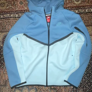  Nike tech fleece  - Nike hoodie i två nyanser av blått med svart dragkedja och huva. Snygg sportig look med logga på bröstet och ärmficka med dragkedja. Tillverkad i mjukt syntetmaterial, perfekt för kyliga dagar. Passar dig som gillar streetwear och sportig stil. Köpt för 1100 och säljer för 700. Pris kan diskuteras