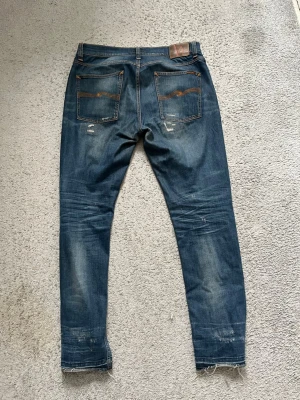 Nudie Jeans blå jeans 34/32 - Säljer ett par blå jeans från Nudie Jeans i storlek 34/32. De har slitningar och trasiga detaljer på benen, klassisk femficksmodell och orange sömmar. Jeansen har en rak passform och är tillverkade i jeansmaterial med råa kanter vid benslut.