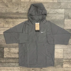 Grå vindjacka nike Miller  - Snygg grå vindjacka från Nike med huva och dragkedja framtill. Jackan har en minimalistisk look med diskret Nike-logga på bröstet och praktiska sidofickor. Perfekt för blåsiga dagar och enkel att matcha med olika outfits.