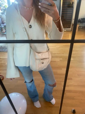 Vit axelväska med dragkedja - Säljer en vit axelväska i skinnimitation med justerbar rem. Väskan har en praktisk framficka med dragkedja och en rund metallknapp. Perfekt storlek för det viktigaste och enkel att matcha till olika outfits.