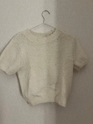 Stickad topp från Gina Tricot - Mjuk och bekväm stickad topp från Gina Tricot i storlek S. Kortärmad modell med rund halsringning och ribbade avslut vid hals, ärmar och nederkant. Färgen är offwhite/ljusbeige. I fint begagnat skick utan tydliga skador eller fläckar.