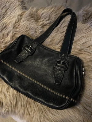 Svart axelväska från Lacoste i skinn - Snygg svart axelväska från Lacoste i äkta skinn med grov struktur. Väskan har dubbla axelband med spänndetaljer, kontrastsömmar och dragkedja upptill. Insidan är ljus med flera fack och en innerficka med dragkedja. Klassisk krokodil-logga på framsidan.