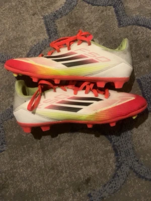 Adidas vita och röda fotbollsskor - Säljer ett par Adidas fotbollsskor i vitt med röda och gula detaljer samt svarta ränder på sidorna. Skorna har snörning, platt sula med fasta dobbar och insida i ljusgrönt meshmaterial. Perfekta för dig som vill sticka ut på planen!
