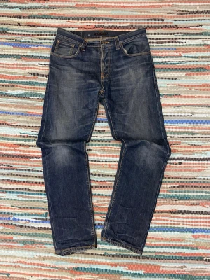 Mörkblå Nudie Jeans straight fit - Säljer ett par mörkblå jeans från Nudie Jeans med klassisk femficksdesign och kontrastsömmar. Modellen har raka ben och snygg slitning på både fram- och baksida samt slitna detaljer vid bensluten. Perfekta för dig som gillar en avslappnad och tidlös look.