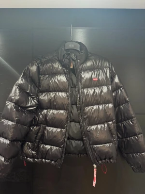 Svart pufferjacka från Levi's M - Säljer en svart pufferjacka från Levi's i storlek M. Jackan har quiltad design, hög krage och dragkedja framtill. Tillverkad i glansig polyester med elastisk nederkant och praktiska fickor. Klassisk Levi's-logga på bröstet och snygga röda detaljer.