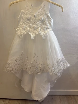 Vit långklänning med spets och pärlor - Elegant vit långklänning med vacker spets och broderade blommor på överdelen. Kjolen är i flera lager tyll med glittriga paljettdetaljer och spetskant. Dekorativa tygrosor och pärlor framtill ger en extra lyxig känsla. Perfekt för speciella tillfällen.