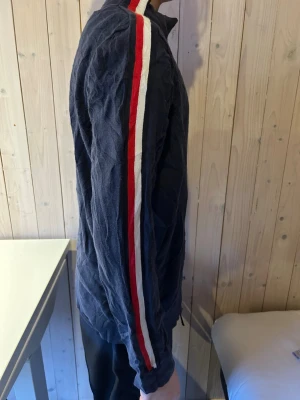 Marinblå polotröja Tommy Hilfiger - Snygg marinblå polotröja från Tommy Hilfiger med röda och vita ränder längs ärmarna. Tröjan har hög krage och diskret logga på bröstet. Tillverkad i mjuk bomullsmix och har lång ärm. Perfekt för dig som gillar sportig och stilren look.Finns dock ingen tag i förutom lappen på bilden