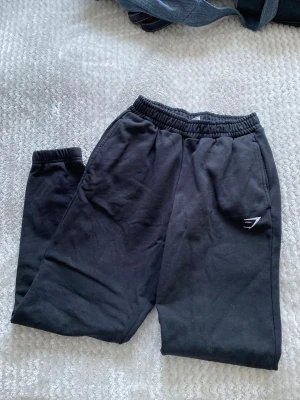 Svarta mjukisbyxor från Gymshark XXS - Säljer ett par svarta mjukisbyxor från Gymshark i storlek XXS. Byxorna har elastisk midja och muddar vid bensluten, samt en liten Gymshark-logga på ena benet. Perfekta för chill eller träning.