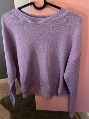 Ljuslila stickad tröja - En enkel och stilren ljuslila stickad tröja med rund halsringning och ribbade muddar. Tröjan har lång ärm och är perfekt för dig som gillar en mjuk och bekväm vardagslook. Passar till jeans eller kjol för en avslappnad stil.