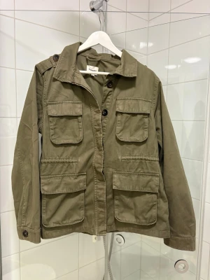 Olivgrön jacka - Säljer en olivgrön jacka från Kappahl i storlek XS. Jackan har fyra stora fickor framtill, axelklaffar med knapp och klassisk krage. Tillverkad i bomull och stängs med knappar. 