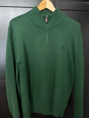 Grön stickad tröja Polo Ralph Lauren - Mörkgrön stickad tröja från Polo Ralph Lauren med halv dragkedja och ribbad hög krage. Klassisk broderad logga på bröstet och ribbade muddar. Perfekt för kyliga dagar och riktigt snygg till jeans.
