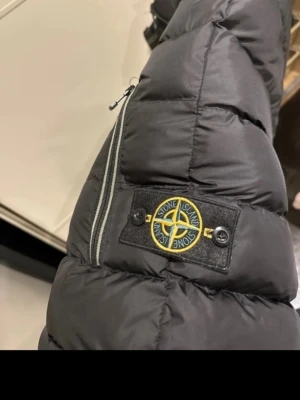 Svart dunväst från Stone Island - Säljer en svart dunväst med hög krage och logga på kragen samt ärmen. Västen har två fickor med dragkedja och quiltad design. Perfekt för lager-på-lager och streetwear-stil. Large men liten i storleken så passar även M 