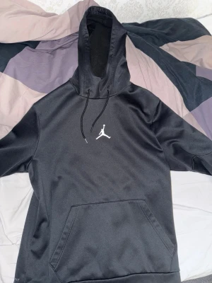 Jordan hoodie - Svart hoodie från Jordan med klassisk vit Jumpman-logga på bröstet. Tröjan har huva med snörning och en stor magficka framtill. Tillverkad i mjukt och lite glansigt polyestermaterial, perfekt för chill eller sport. Liten defekt på ena änden av snöret. Fraktas endast och köparen står för frakt.