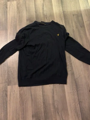 Mörkblå stickad tröja från Lyle & Scott - Svart stickad tröja från Lyle & Scott med rund hals och diskret gul logga på bröstet. Tröjan har lång ärm och klassisk passform, perfekt för lager-på-lager under kyliga dagar.