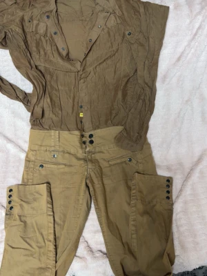 Bruna cargopants från G-Star RAW - Säljer ett par bruna cargopants från G-Star RAW med hög midja och coola knappdetaljer både fram och vid bensluten. Byxorna har flera fickor och är gjorda i bomullsmaterial som känns skönt mot huden. Perfekt för dig som gillar streetstyle och funktionella detaljer.