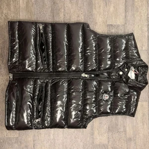 Svart glansig dunväst från Moncler - Snygg svart glansig dunväst från Moncler med dragkedja och hög krage. Västen har två praktiska fickor med dragkedjor framtill och en stilren design som passar perfekt för kyligare dagar.