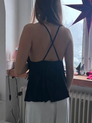 Svart ribbad linnetopp med korsade band - Säljer en svart ribbad linnetopp med tunna axelband som korsas i ryggen. Toppen har en rak, något lös passform och ärmlös design. Materialet är mjukt och stretchigt, perfekt för sommardagar eller lager på lager. Enkel men stilren look som passar till mycket.