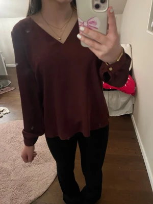 Vinröd/brun blus  - Jag säljer denna jättefina blus som har en v-ringning med lång ärm och en liten gulddetalj i ärmens ände 