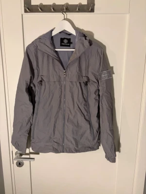 Grå vindjacka från Stone Island - Säljer en grå vindjacka från Stone Island med ikonisk patch på ärmen. Jackan har huva, meshfoder och två dragkedjeförsedda fickor framtill. Tillverkad i lätt polyester och har hel dragkedja. Perfekt för dig som vill ha en clean och stilren look. Väldigt sällsynt jacka 