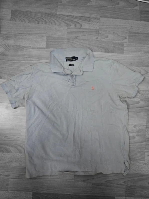 Ljusblå piké från Polo Ralph Lauren XL - Ljusblå pikétröja från Polo Ralph Lauren i custom fit. Klassisk krage, korta ärmar och den ikoniska broderade loggan i rött på bröstet. Tillverkad i mjuk bomull som är skön att bära. Perfekt till jeans eller shorts för en clean look.