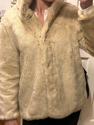 Beige pälsjacka med huva - Säljer en fluffig beige pälsjacka med huva och knäppning framtill. Jackan är helfärgad och har en mjuk, gosig känsla som passar perfekt när det är kallt ute. Långärmad modell med en lite loose passform. Perfekt för dig som vill ha en mysig och snygg look.